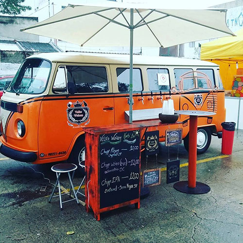 Kombi com a metade de baixo cor de laranja e a metade de cima bege claro, divididas por duas faixas horizontais pretas que, na frente do veículo, formam um desenho em V. Na porta do motorista, logomarca em preto e branco. Na lateral, lado a lado logo abaixo das janelas, 5 torneiras de chopeira e pequena bancada de apoio para copos. Ao lado, toldo em tecido branco no formato de um grande e alto guarda-sol. Embaixo, 2 banquetas, bancada com cardápio escrito sobre quadro negro na lateral, pequena mesa redonda bem alta e uma lixeira vermelha.